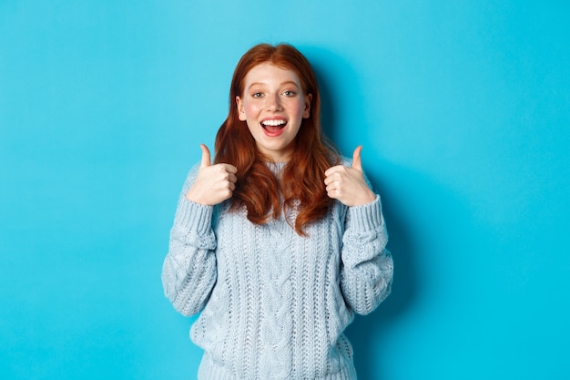 Supportive redhead girl in sweater, showing thumbs-up and looking amazed, praising good choice, standing over blue background.