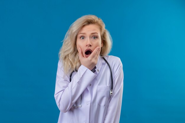 Suprised doctor young girl wearing stethoscope in medical gown put her hand on jaw on blue background