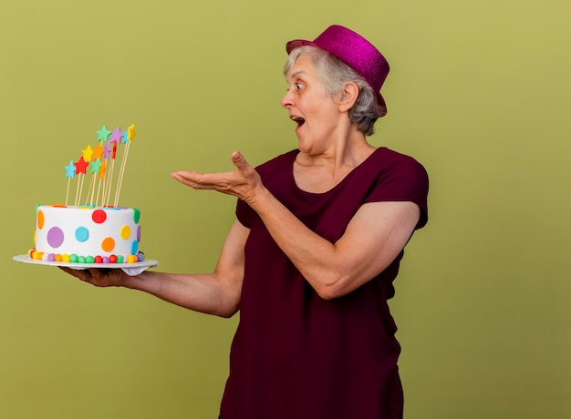 Удивленная пожилая женщина в праздничной шляпе держит и указывает на праздничный торт, изолированный на оливково-зеленой стене с копией пространства