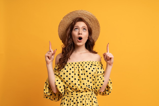 Suprised red-haired woman  posing in yellow dress with sleeves pointing up by fingers on yellow. Summer mood.