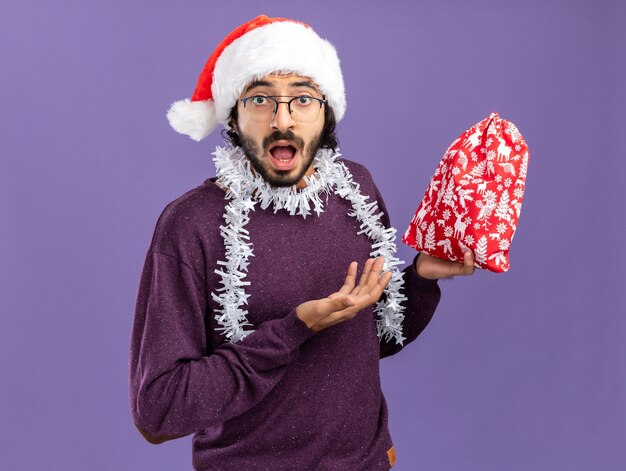 Suprised young handsome guy wearing christmas hat with garland on neck holding and points at christmas bag isolated on blue background