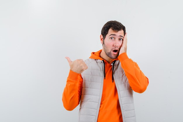 The suprised young man is putting ona hand on cheek and pointing to left with other on white background