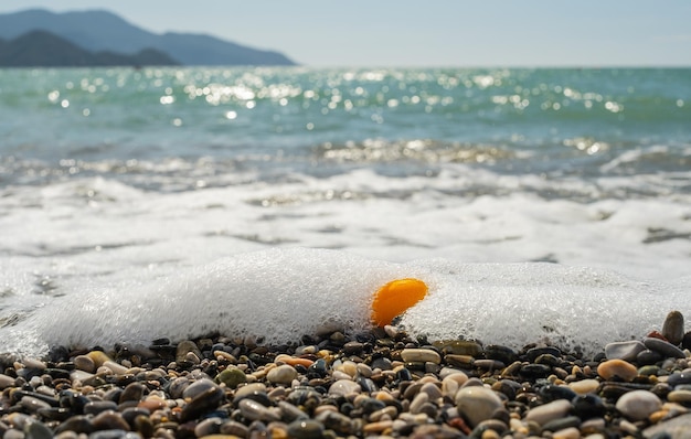 Серфинг на галечном пляже ярко-оранжевый плод кумквата в морской пене, переносимый волной на пляж, избирательный фокус на море в яркий солнечный день, время отпуска или идея для фона