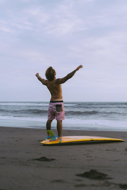 サーファーの男が砂浜をボードを持って歩きます。ビーチでハンサムな若い男。ウォータースポーツ。健康的なアクティブなライフスタイル。サーフィン。夏休み。エクストリームスポーツ。