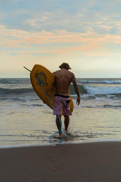 A surfer man walks with a board on a sandy beach. Handsome young man on the beach. water sports. Healthy active lifestyle. Surfing. Summer vacation. Extreme sport.