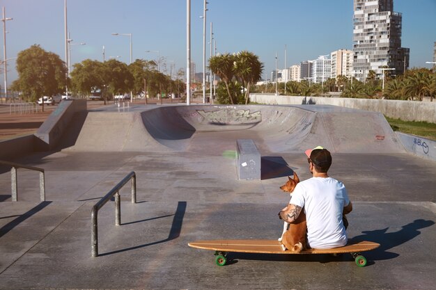 Surfer sits on a longboard in a city skatepark hugging his brown and white basenji dog, looking on city landscape, shot from the back