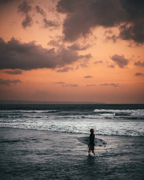 A surfer wearing surfing swimsuit holding a surfboard standing at the seashore during sunset