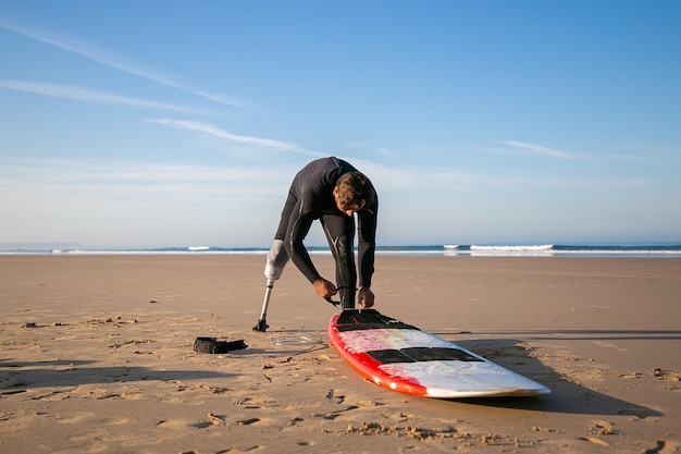 Surfer wearing wetsuit and artificial limb, tying board to his ankle on sand