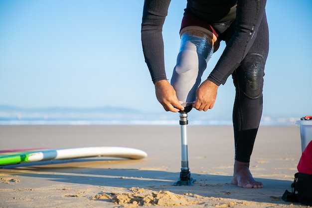 Free photo surfer wearing wetsuit, standing by surfboard on sand and adjusting artificial limb taped to leg