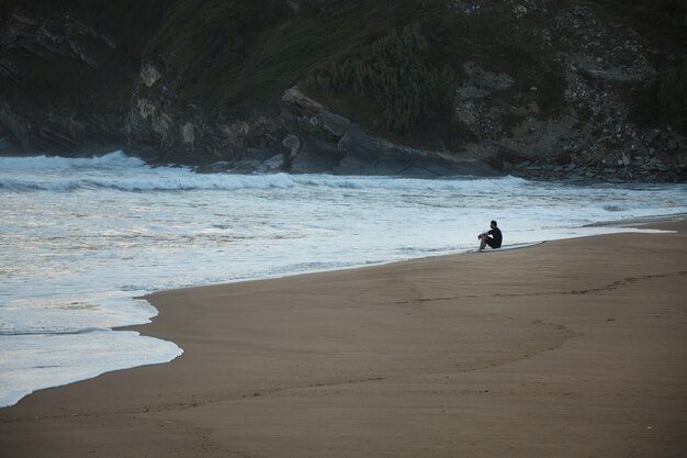 Surfer in wetsuit sitting at the edge of a sandy beach under a green and rocky hill in the evening