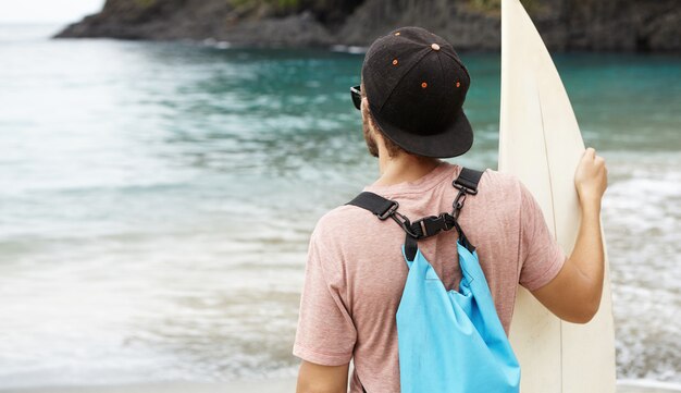 Surfer with surfboard relaxing on beach after training, watching other surfers practicing new tricks. Back shot of Caucasian man enjoying beautiful view in front of him