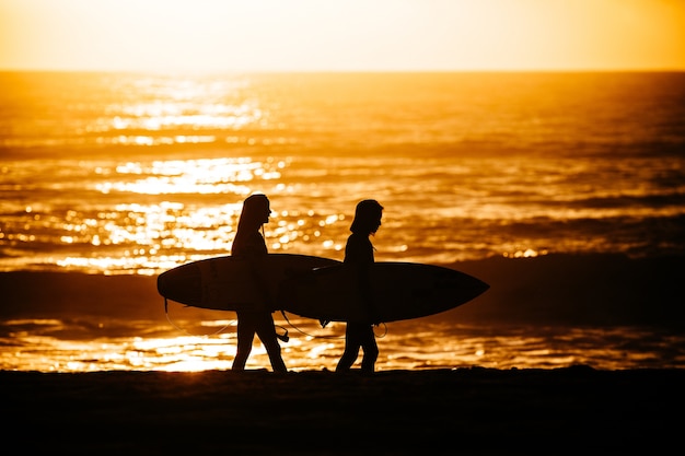 Surfers walking after an exhausting surfing session against a dazzling sunset background