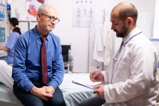 Surgeon wearing stethoscope discussing treatment in examination room with senior man