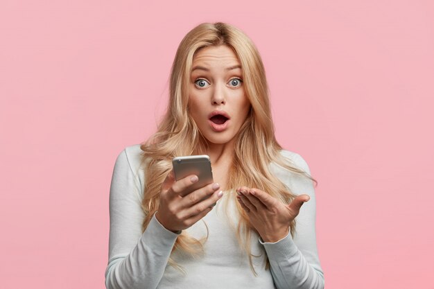 Surpised businesswoman reads unexpected news about startup, checks email on modern cell phone, looks worried as recives message from boss, isolated over pink wall. Omg and surprisment concept