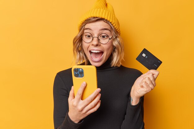 Surpised happy woman wears hat and casual black turtleneck looks at smartphone app screen holds credit card uses bank account for paying online isolated over yellow background gets cashback