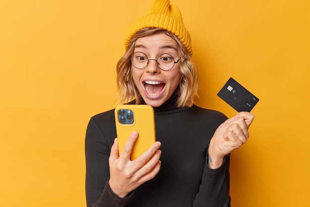 Surpised happy woman wears hat and casual black turtleneck looks at smartphone app screen holds credit card uses bank account for paying online isolated over yellow background gets cashback