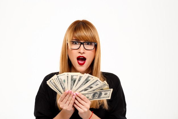 Surpised successful businesswoman in glasses holding money over white wall