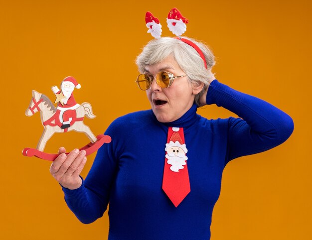 Surpried elderly woman in sun glasses with santa headband and santa tie holding and looking at santa on rocking horse decoration isolated on orange background with copy space