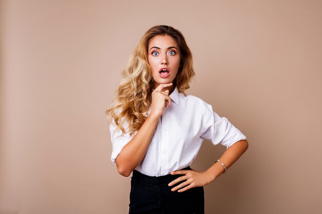 Surprise face. Blond woman in casual outfit standing over beige wall. Exited girl looking.