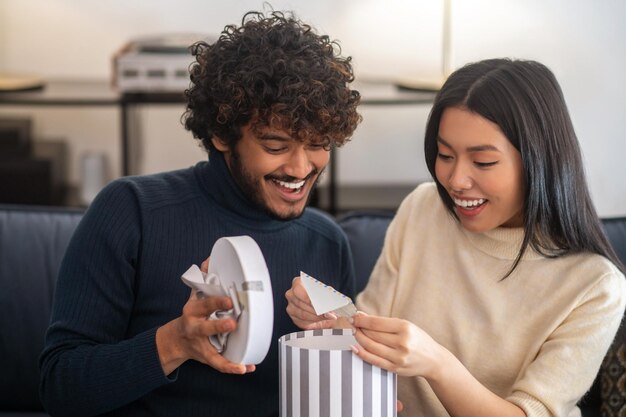 Surprise, joy. Smiling attentive curly-haired indian man opening gift light box and pleasantly surprised beautiful asian woman pulling out letter sitting on sofa