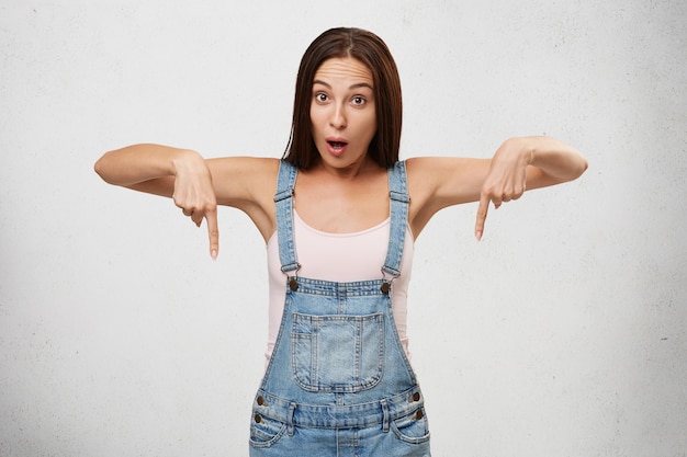 Foto gratuita concetto di sorpresa, shock e stupore. bella donna scioccata in tuta di jeans, che punta verso il basso entrambi gli indici, indicando qualcosa di sorprendente