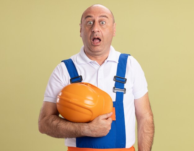 Surprised adult builder man in uniform holds safety helmet isolated on olive green wall