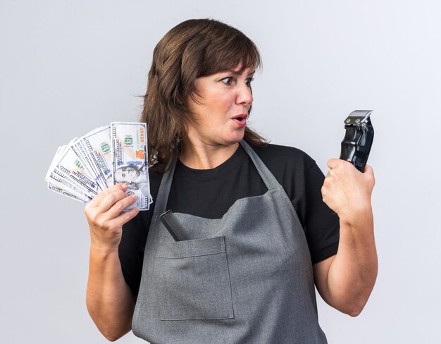 Surprised adult caucasian female barber in uniform holding money and looking at hair clipper isolated on white wall with copy space
