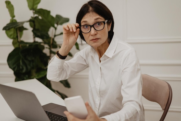 La donna caucasica adulta sorpresa guarda in modo sorprendente la fotocamera mentre si regola gli occhiali mentre è seduto a tavola con il laptop concetto di emozione espressione facciale