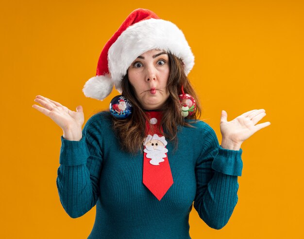 Surprised adult caucasian woman with santa hat and santa tie holds glass ball ornaments on ears making fish mouth and keeping hands open isolated on orange background with copy space