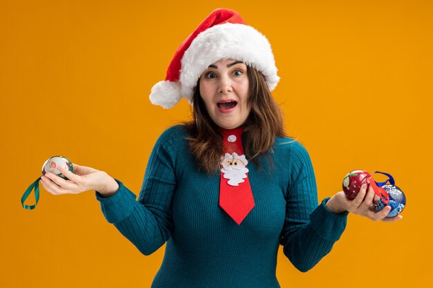 Surprised adult caucasian woman with santa hat and santa tie holds glass ball ornaments 