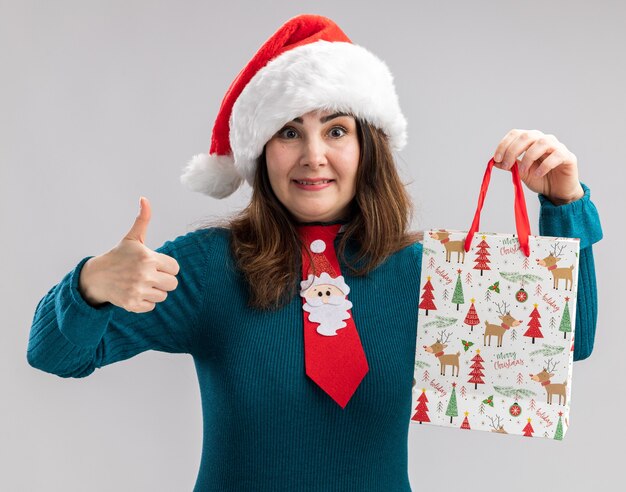 Surprised adult caucasian woman with santa hat and santa tie holds paper gift box and thumbs up isolated on white background with copy space