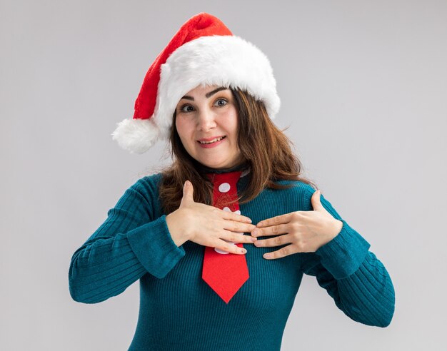 Surprised adult caucasian woman with santa hat and santa tie puts hands on chest isolated on white wall with copy space