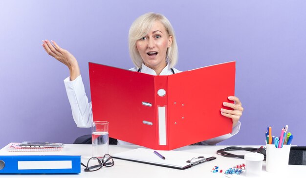 surprised adult female doctor in medical robe with stethoscope sitting at desk with office tools holding file folder isolated on purple wall with copy space