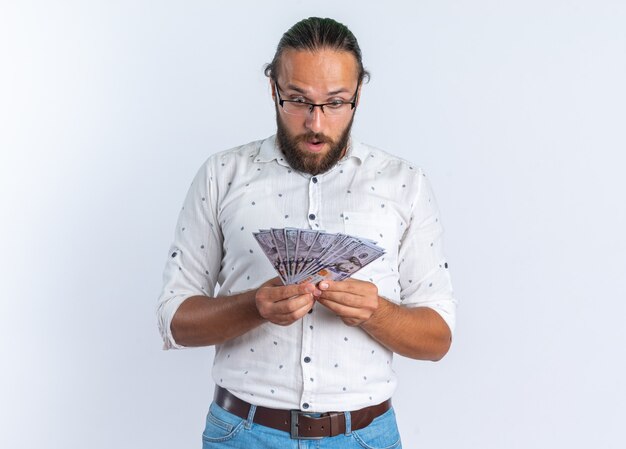 Foto gratuita sorpreso adulto bell'uomo con gli occhiali che tiene e guarda i soldi isolati sul muro bianco con copia spazio
