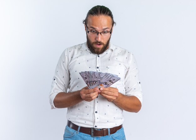 Surprised adult handsome man wearing glasses holding and looking at money isolated on white wall with copy space