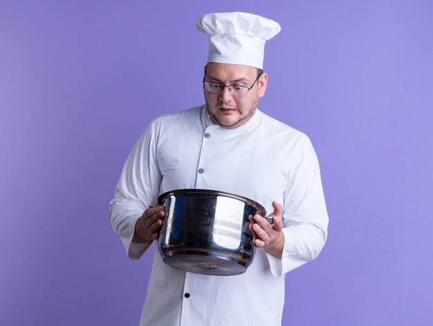 удивленный взрослый мужчина-повар в униформе шеф-повара и очках держит горшок, глядя внутрь него, изолированного на фиолетовой стене с копией пространства
