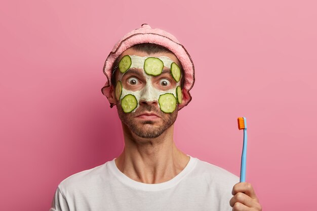 Surprised adult man cares about skin, applies facial mask and slices of cucumber, holds toothbrush