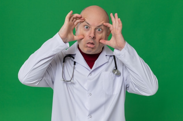 Удивленный взрослый мужчина в униформе врача со стетоскопом, открыв глаза руками и глядя