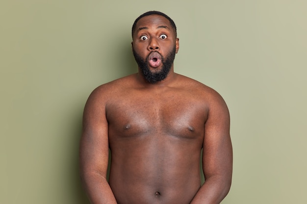 Free photo surprised adult man with beard stands shirtless stares at front keeps mouth widely opened reacts on something unexpected and shocked isolated over studio wall of khaki color