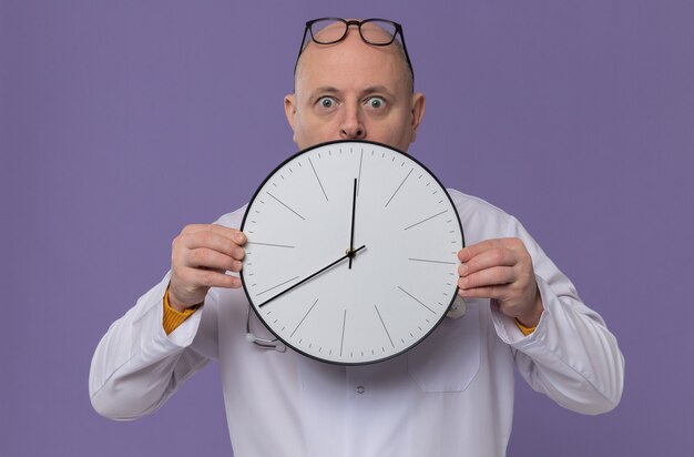 Surprised adult man with glasses in doctor uniform with stethoscope holding clock 