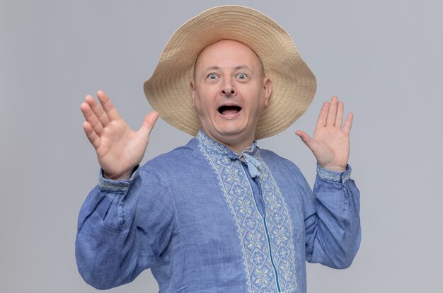 Surprised adult man with straw hat and in blue shirt standing with raised hands 