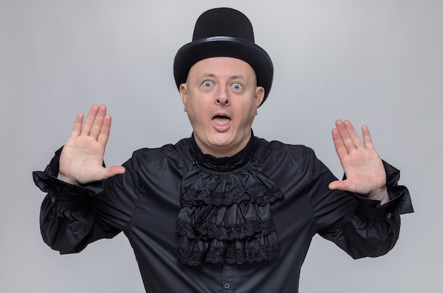 Foto gratuita uomo adulto sorpreso con cappello a cilindro e camicia gotica nera in piedi con le mani alzate