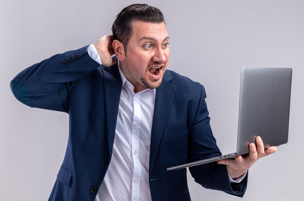 Surprised adult slavic businessman holding and looking at laptop isolated on white wall with copy space