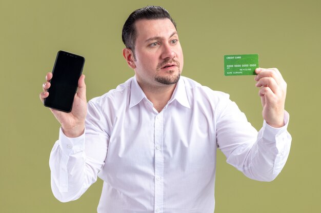 Удивленный взрослый славянский бизнесмен держит телефон и смотрит на кредитную карту