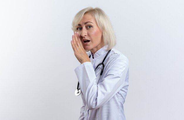 Surprised adult slavic female doctor in medical robe with stethoscope holding hand close to her mouth isolated on white wall with copy space