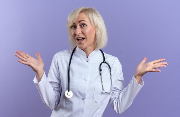 Surprised adult slavic female doctor in medical robe with stethoscope keeping hands open isolated on purple background with copy space