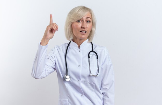 Surprised adult slavic female doctor in medical robe with stethoscope pointing up isolated on white background with copy space