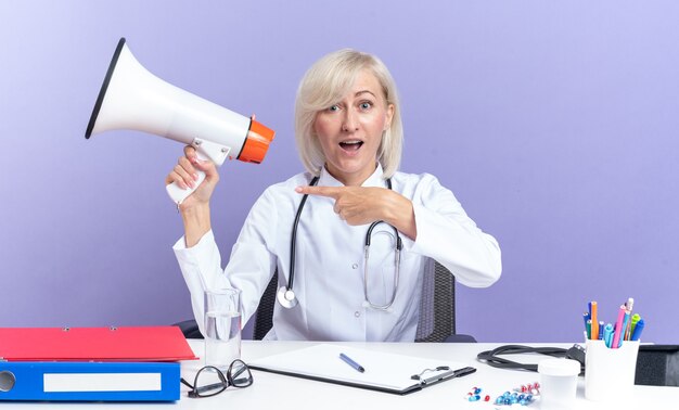 Surprised adult slavic female doctor in medical robe with stethoscope sitting at desk with office tools holding and pointing at loud speaker isolated on purple background with copy space