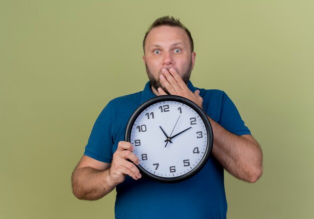 Surprised adult slavic man holding clock putting hand on mouth looking 