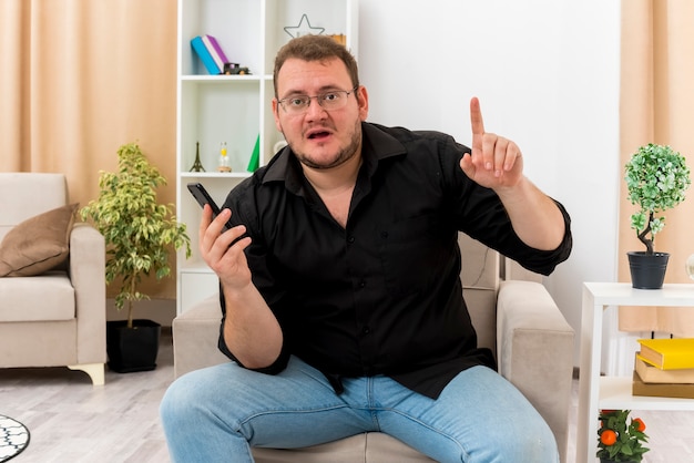 Surprised adult slavic man in optical glasses sits on armchair holding phone and pointing up inside the living room
